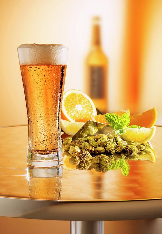 Freshly Drawn Beer In A Glass On A Metal Table In Front Of An Arrangement Of Hops, Lemons And A Beer Bottle Photograph by Julian Winkhaus