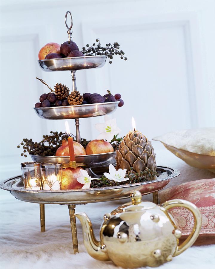 Fruit, Artificial Flowers And Candles Arranged On Shiny Silver Cake Stand Behind Shiny Gold Teapot Photograph by Matteo Manduzio