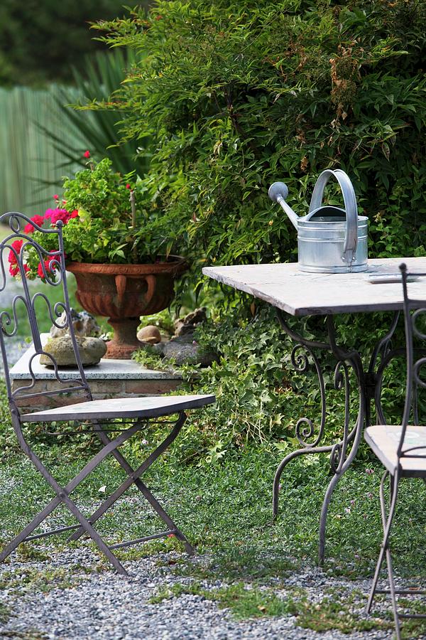 Garden Table With Watering Can And Garden Chair In Front Of Bush And Planted Urn Photograph by Ulrike Schmid