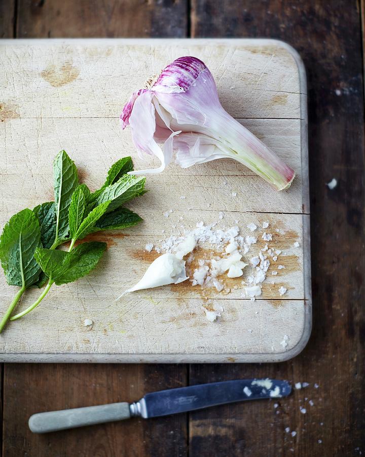 Garlic, Sea Salt And Mint Leaves On A Wooden Chopping Board Photograph by Tom Regester