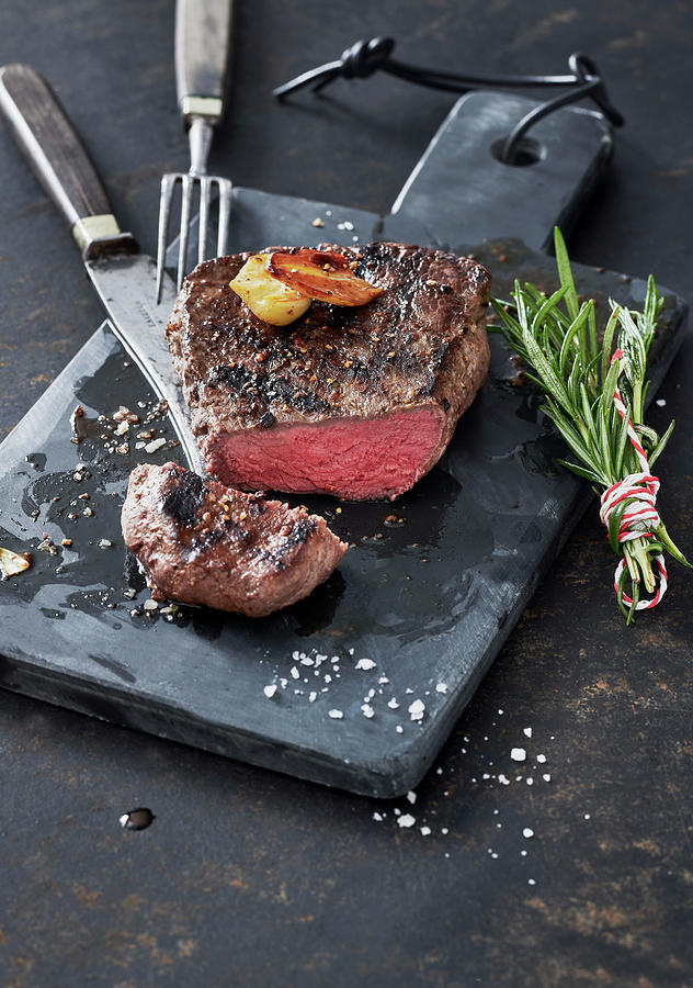 Grilled Steak With Garlic And A Bundle Of Rosemary On A Slate Board Photograph by Stefan Schulte-ladbeck