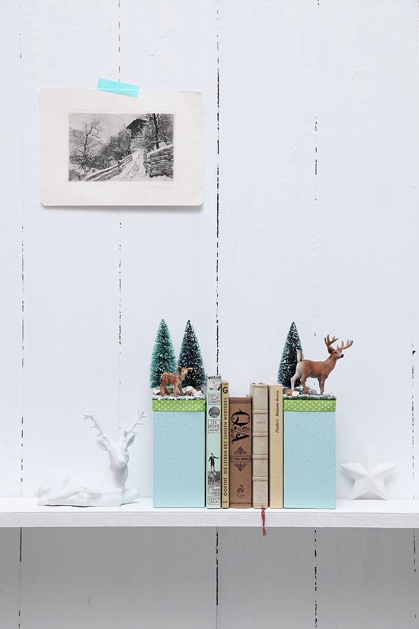 Hand-crafted Bookends With Winter Motifs Decorated With Miniature Deer And Tiny Fir Trees On Shelf On White, Wooden Wall Photograph by Thordis Rggeberg