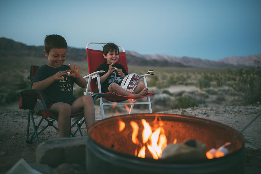 Joshua Tree National Park Photograph - Happy Brothers Eating Marshmallows While Sitting On Chairs At Campsite During Dusk by Cavan Images