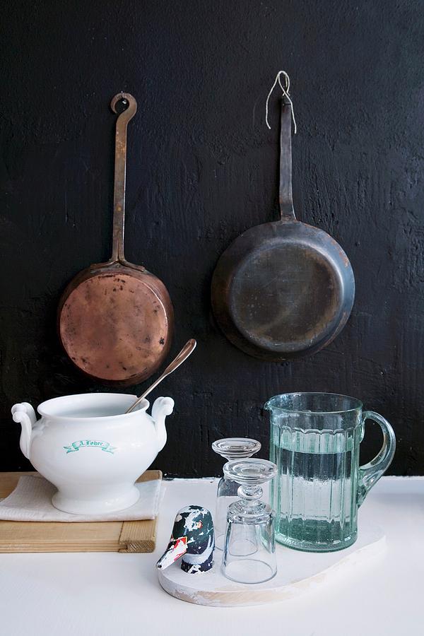 Jug Of Water, Glasses And Soup Tureen In Front Of Vintage Pans Hanging On Black Wall Photograph by Sandra Eckhardt