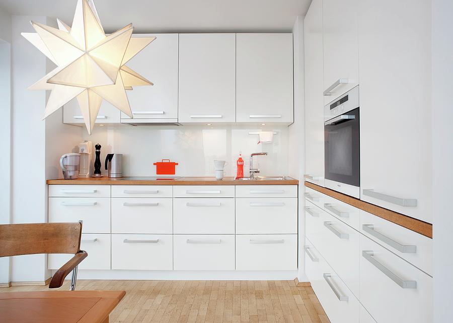 Kitchen With White Furnishings, Wooden Worksurfaces, Fitted Cooker And Star-shaped Lamp Photograph by Hans Gerlach