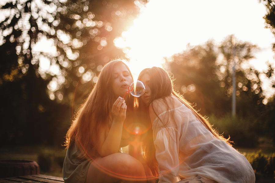 Lesbian Couple Blowing Bubbles While Sitting In Park During Summer