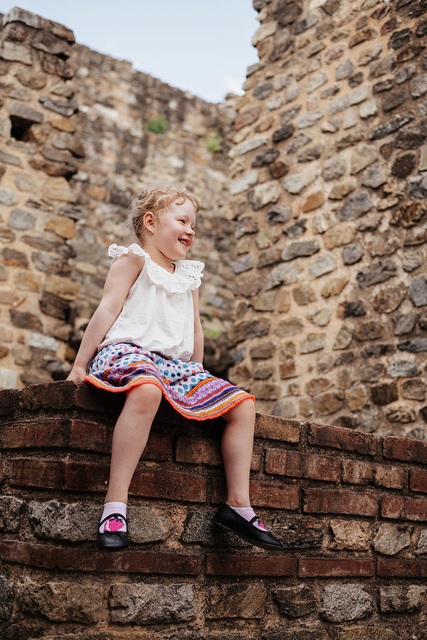 Architecture Photograph - Low Angle View Of Cute Smiling Girl With Blond Hair Sitting Against Old Brick Wall by Cavan Images