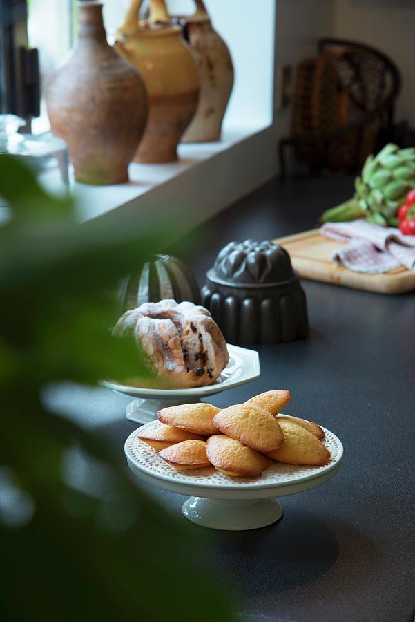 Madeleines And Bundt Cake On Cake Stands Next To Windowsill In Country-house Kitchen Photograph by Christophe Madamour