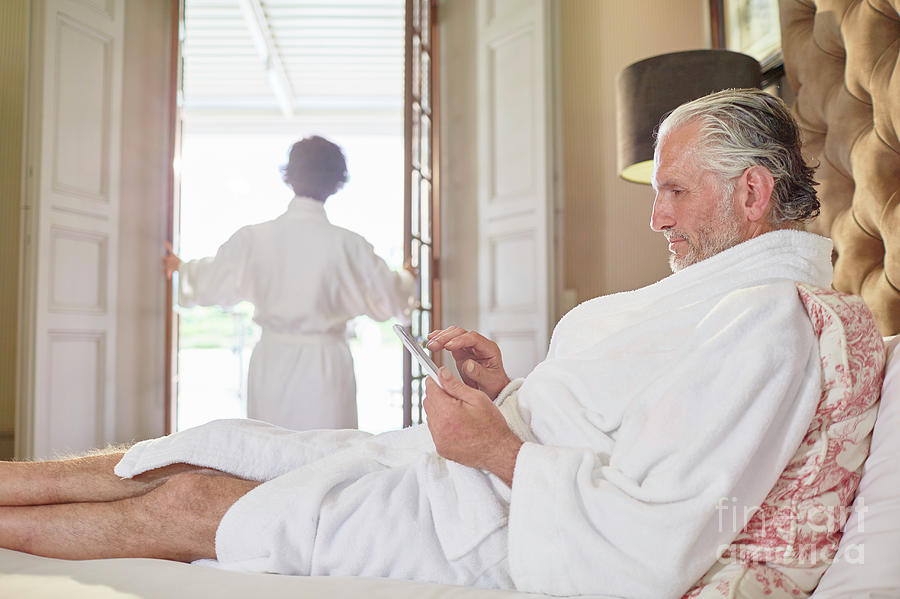 Mature Couple In Bathrobes Lying In Bed And Woman Reading Book In Hotel Room Stock Photo