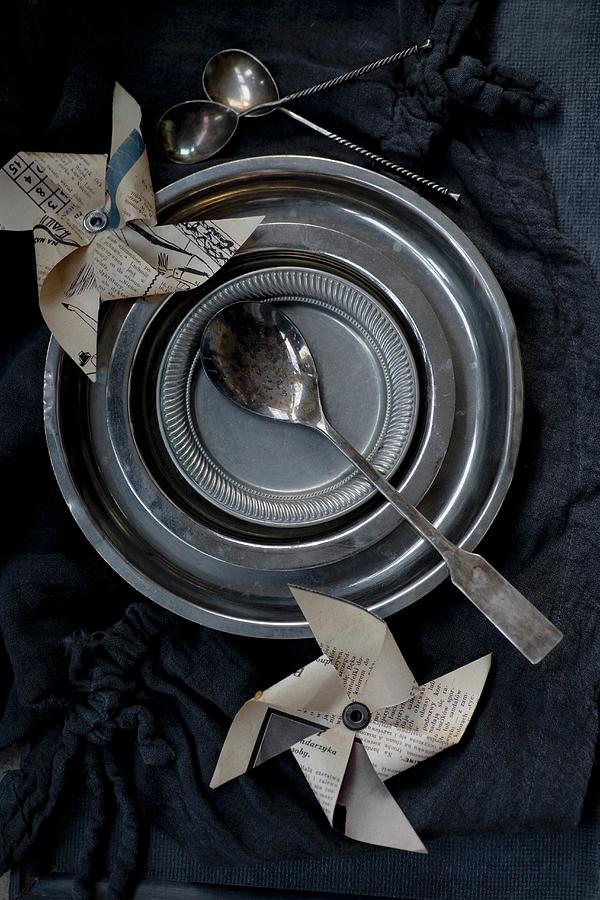 Metal Plates, Various Spoons And Newspaper Windmills On Black Tablecloth Photograph by Alicja Koll