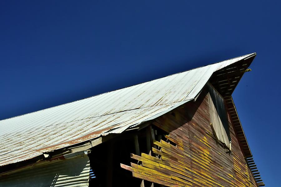 Metal roof peak and hayloft door on exterior of weathered wood shed with yellow patina and blue sky. Photograph by Jerry Sodorff