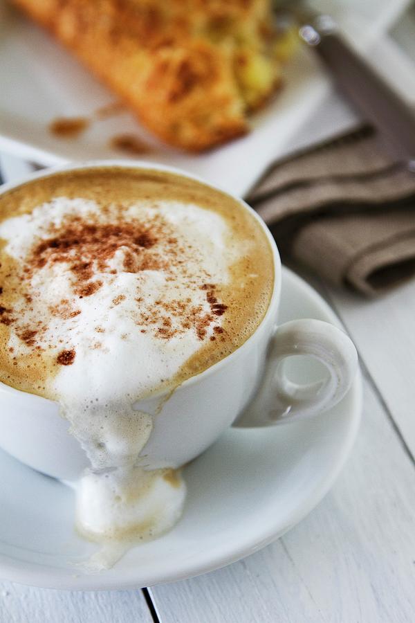 Milk Foam Spilling Out Of A Cappuccino With An Apple Turnover In The Background Photograph by Catja Vedder