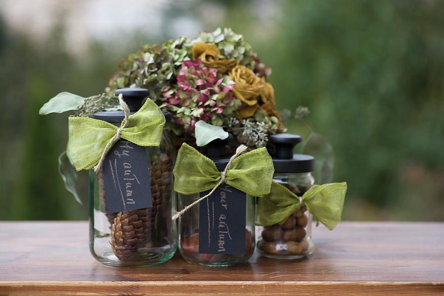 Nuts And Pine Cones In Three Storage Jars With Black Lids And Labels In Front Of Posy Of Dried Flowers Photograph by Alicja Koll