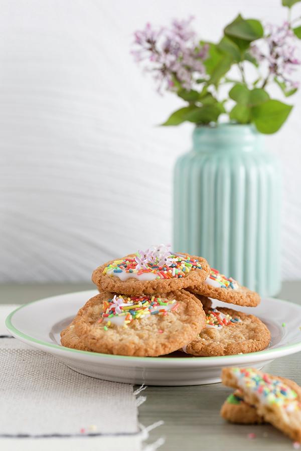 Oat Biscuits With Icing And Sugar Sprinkles With A Vase Of Lilac Flowers In The Background Photograph by Mandy Reschke