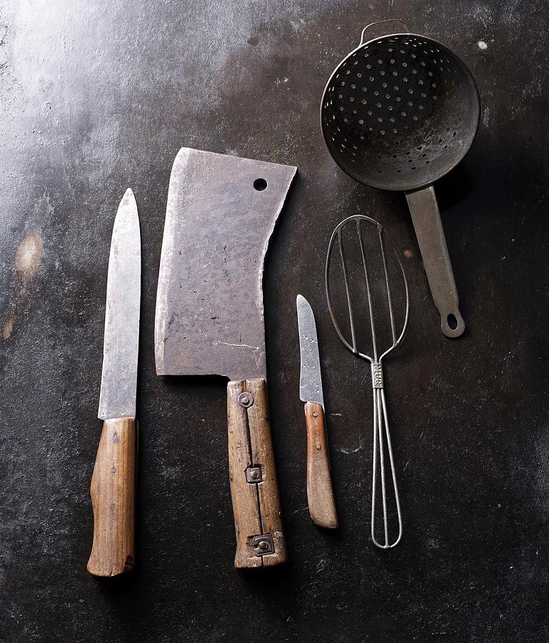 Old Kitchen Utensils On A Dark Background: Meat Cleaver, Wire Spoon, Knives And Colander Photograph by Ludger Rose