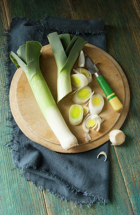 Overhead Shot Of Two Leeks On A Round Wooden Chopping Board Being Sliced By A Retro Wooden Handled Knife And Navy Cloth And Green Wooden Background Photograph by Stacy Grant