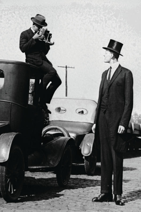 photographer Mounts himself on the roof of a car to shoot a pictures of an exceedingly tall men in a top hat. Painting by 