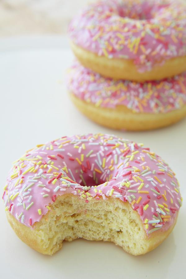 Pink Glazed Dougnuts With Sugar Sprinkles, One With A Bite Taken Out Photograph by Simon Scarboro