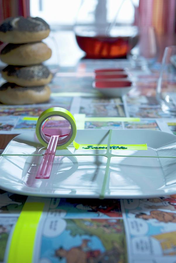Place Setting With Rubber Band Stretched Over Plate And Roll Of Washi Tape As Name Tag On Tablecloth Made From Comic Pages Photograph by Matteo Manduzio