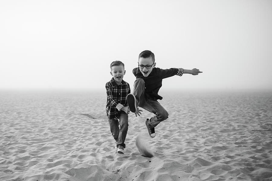 Nature Photograph - Playful Brothers Jumping On Sand At Beach Against Sky During Foggy Weather by Cavan Images