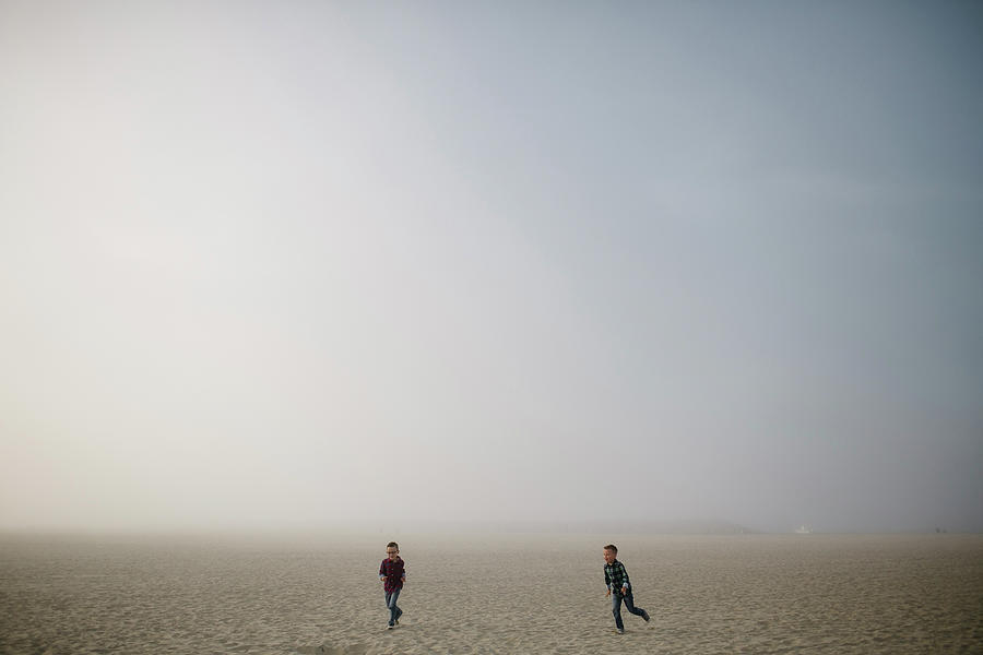 Nature Photograph - Playful Brothers Running On Sand At Beach Against Sky During Foggy Weather by Cavan Images