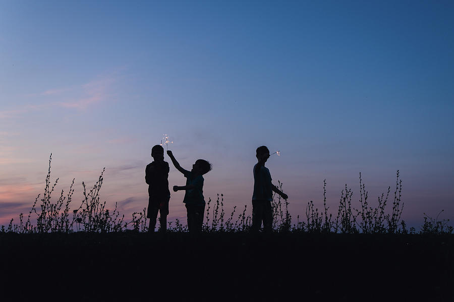 Nature Photograph - Playful Silhouette Brothers Holding Sparklers While Standing On Field Against Sky During Dusk by Cavan Images