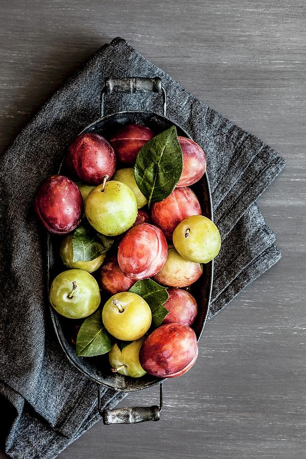 Plums With Leaves In A Metal Bowl On A Grey Cloth Photograph by Sarah Coghill