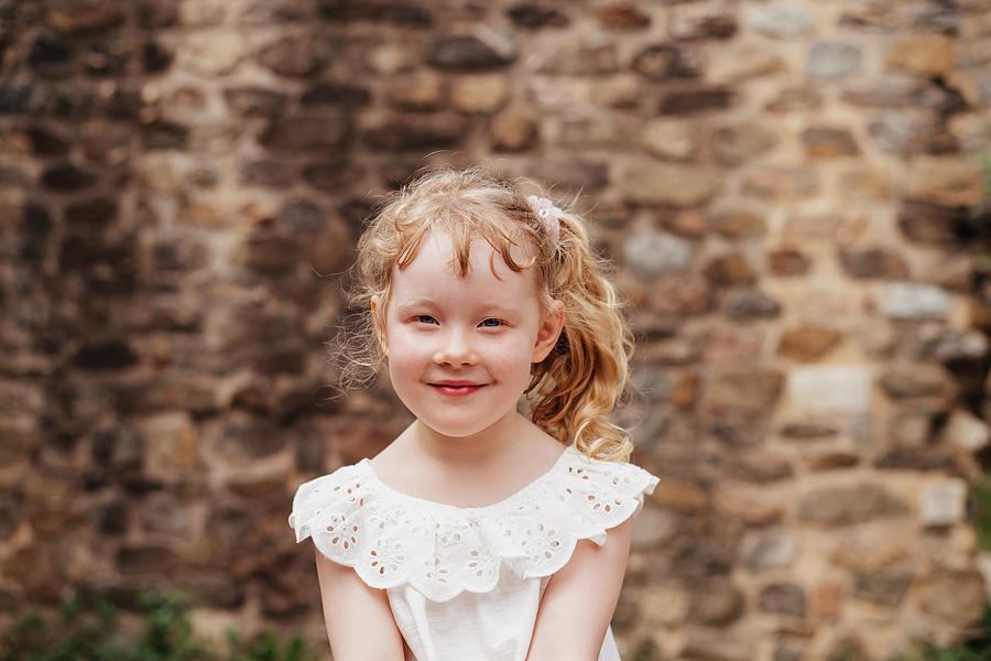 Architecture Photograph - Portrait Of Cute Smiling Girl With Blond Hair Sitting Against Old Brick Wall by Cavan Images