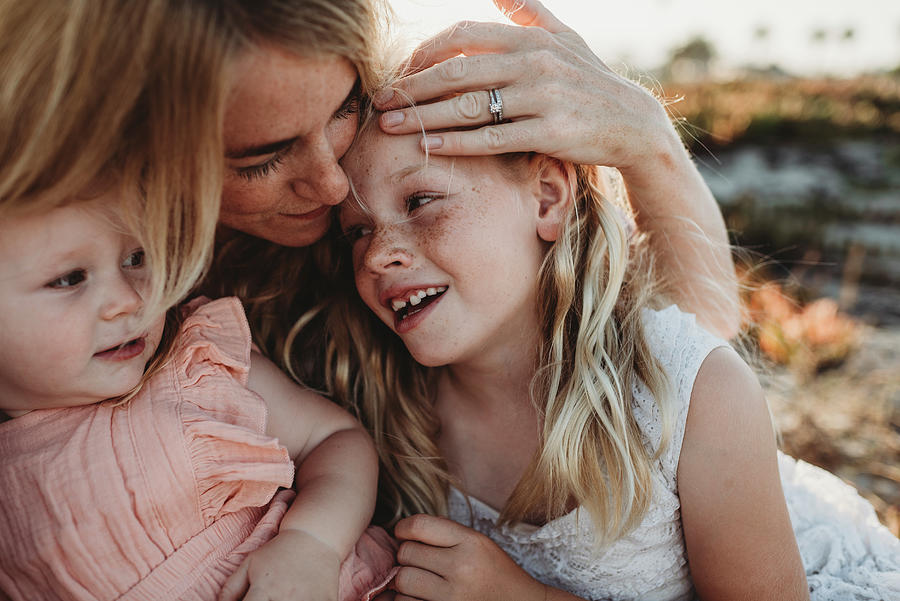 Portrait Of Mother Cuddling With Young Daughters At Beach Sunset 