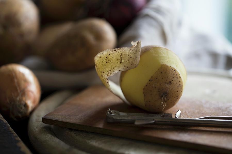 Potatoes And A Peeler On A Wooden Board With Potatoes And Onions In The Background Photograph by Nicole Godt