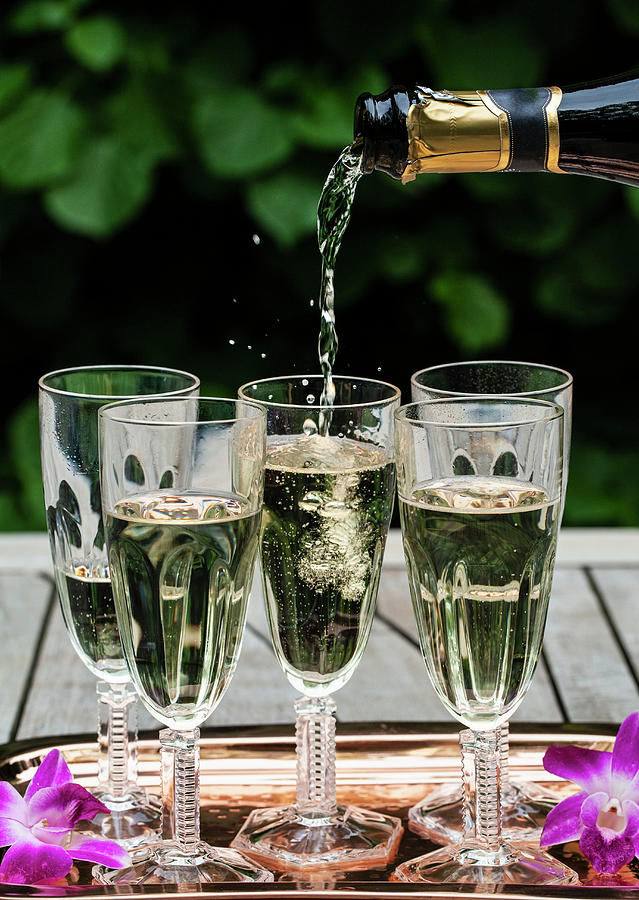 Pouring A Bottle Of Prosecco Into Glass Flutes Lined Up On A Copper Tray Outside, With Purple Orchids, And Greenery Behind Photograph by Ryla Campbell