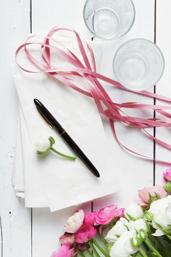Ranunculus, White Paper Bags, Pen, Ribbon And Drinking Glasses On White Wooden Surface Photograph by Catja Vedder