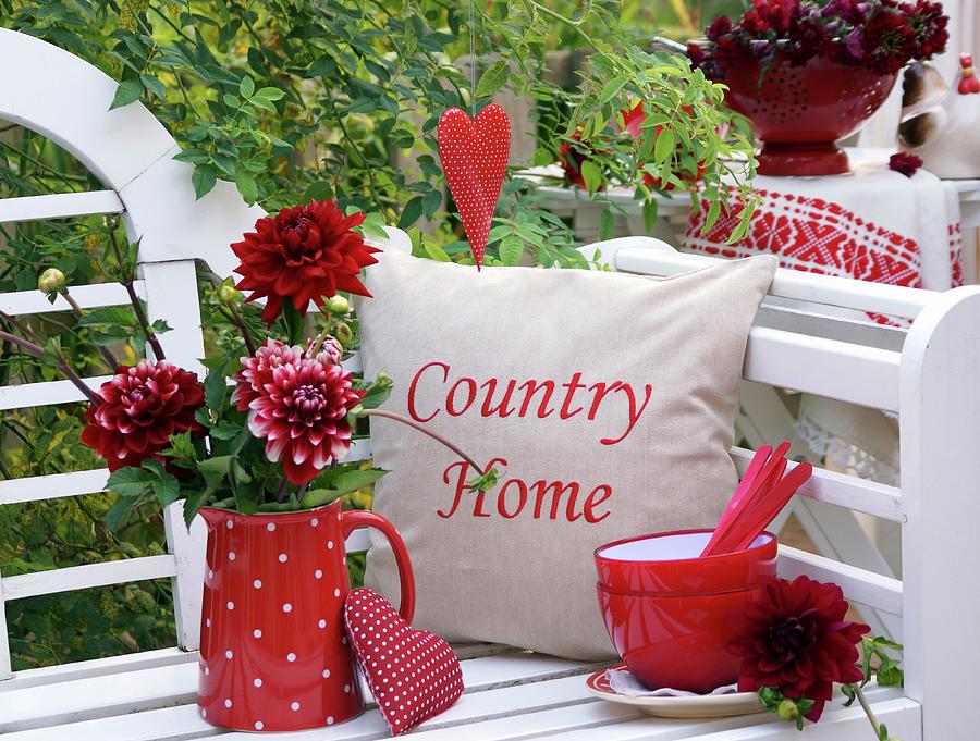 Red Dahlias In Red, Polka-dotted Jug Next To Stack Of Bowls And Cushions On White Garden Bench Photograph by Angelica Linnhoff