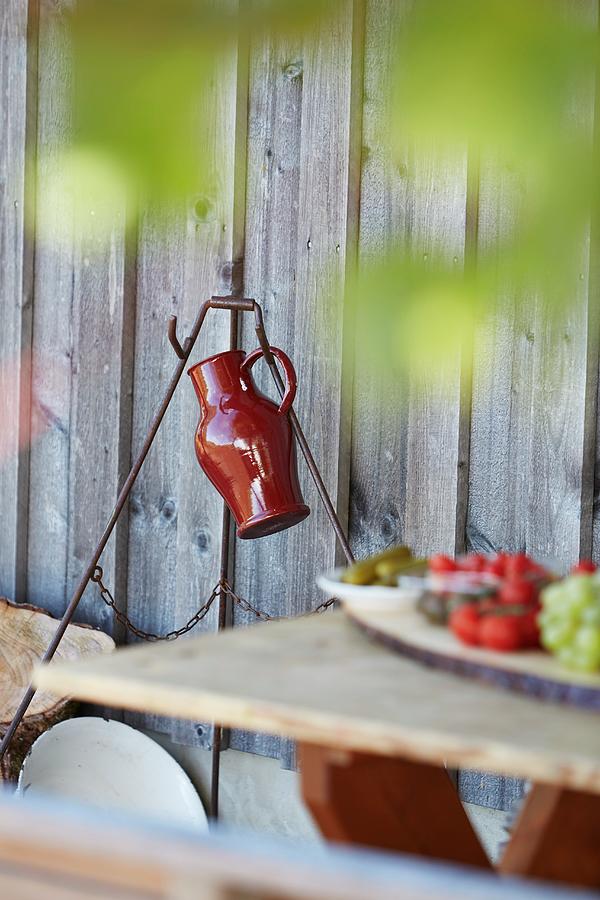 Red Jug Hanging On Metal Frame Against Wooden Wall; Lunch On Table In Blurred Foreground Photograph by Christoph Dpper
