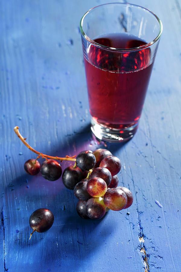 Red Wine Grapes And Grape Juice On A Bllue Wooden Tabletop Photograph by Mandy Reschke