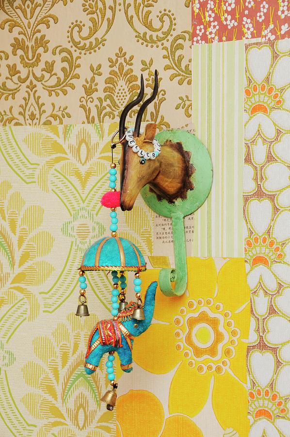 Retro Ornaments In Childs Bedroom; Indian Elephant Mobile Hanging From Tin Coat Hook Shaped Like Hunting Trophy On Patchwork Of 70s Wallpapers Photograph by Revier 51