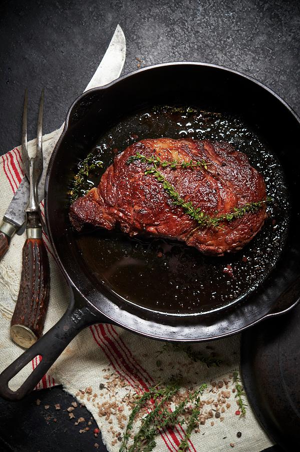 Rib Eye Steak Pan Seared In A Cast Iron Skillet With Thyme; Carving Fork And Knife Photograph by Rannells, Greg