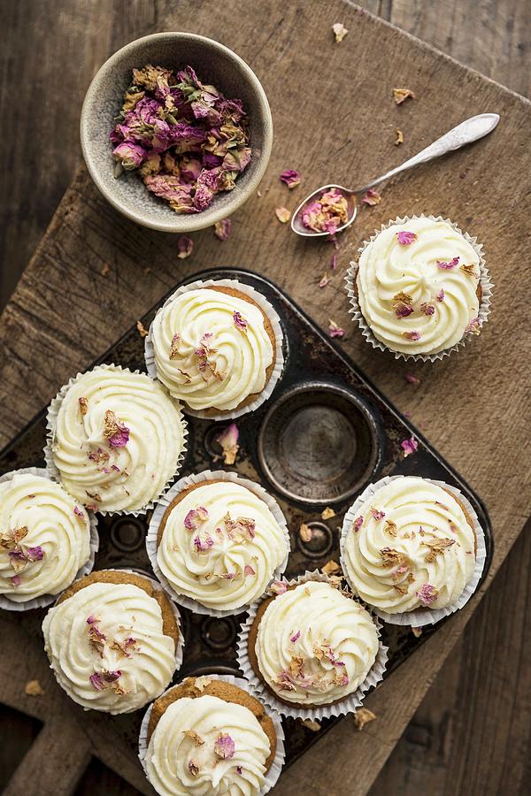 Rose And Lemon Cupcakes With Rosewater And Lemon Frosting In A Rustic Baking Tin Decorated With Dried Edible Rose Petals With Spoon And Bowl Of Rose Petals On A Wooden Board And Table Photograph by Sarah Coghill