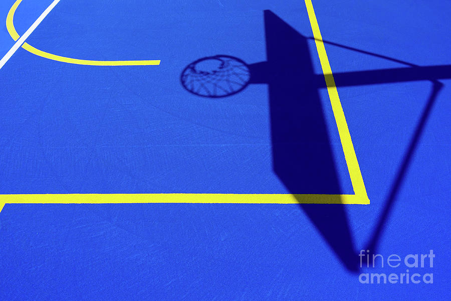 Shadow of a basketball basket on the floor of the court, painted blue and background with lines. Photograph by Joaquin Corbalan