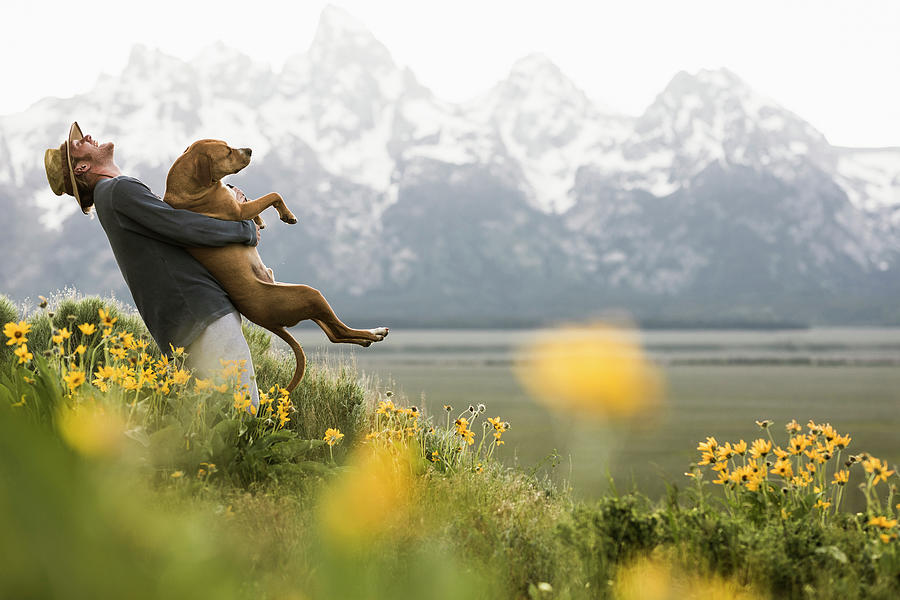 Flower Photograph - Side View Of Happy Man Carrying Dog While Standing Amidst Plants By Lake Against Snowcapped Mountain by Cavan Images