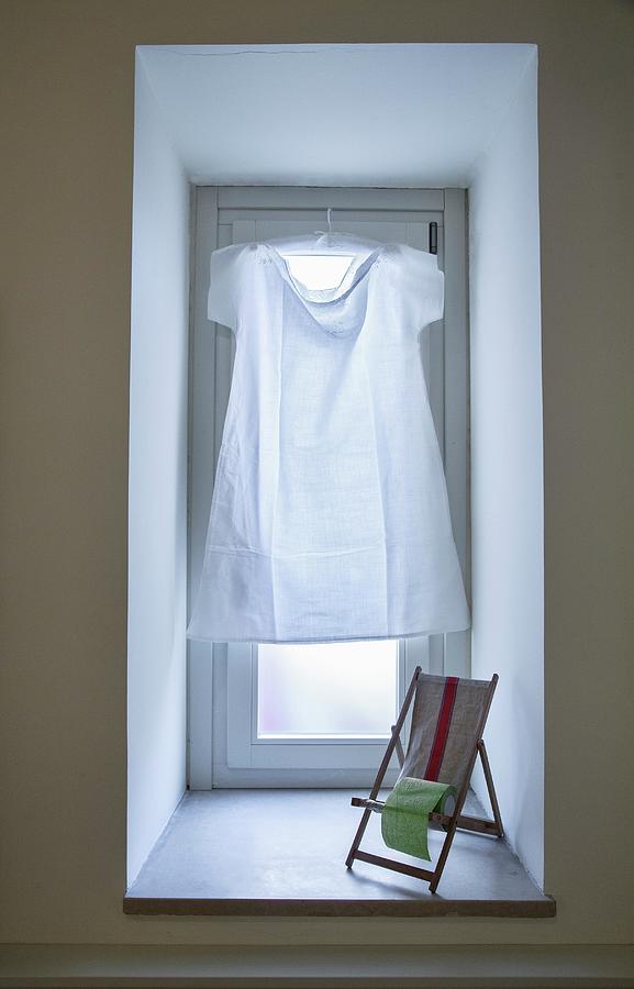 Simple White Dress Hung In Window In Place Of Curtain And Miniature Deckchair On Windowsill Photograph by Laura Rizzi