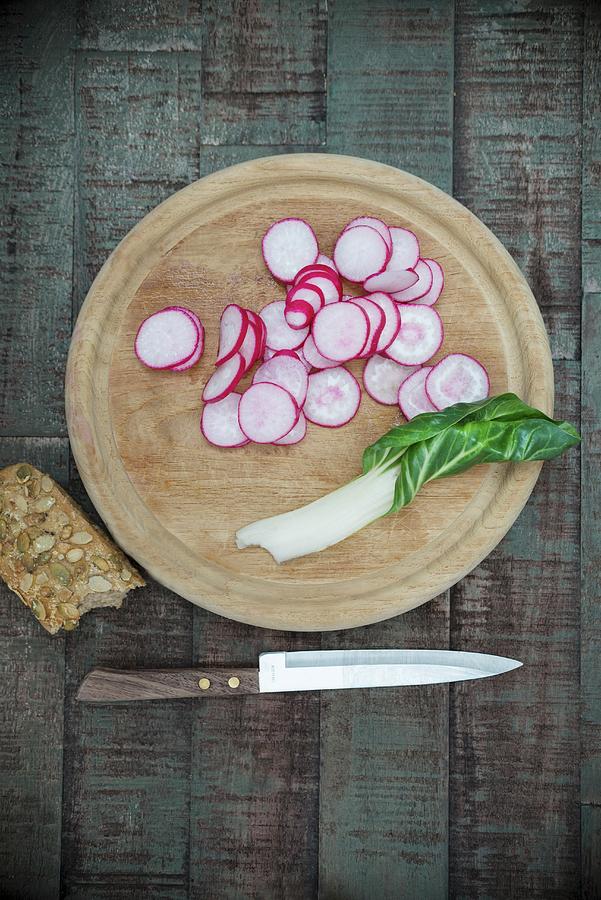 Sliced Radishes And A Chard Leaf On A Wooden Board Next To A Knife And A Sunflower Seed Roll Photograph by Angelika Grossmann