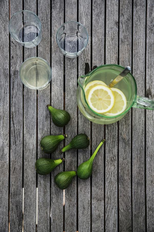 Slices Of Lemon In Glass Vase Of Lemonade, Drinking Glasses And Fresh Green Figs On Wooden Surface Photograph by Jenny Brandt