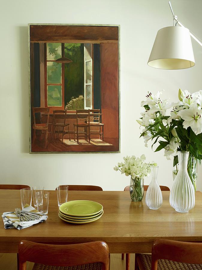 Stacked Plates, Glass Vases And Bouquet Of White Lilies On Oak Dining Table In Front Of Painting On Wall Photograph by Rachael Smith