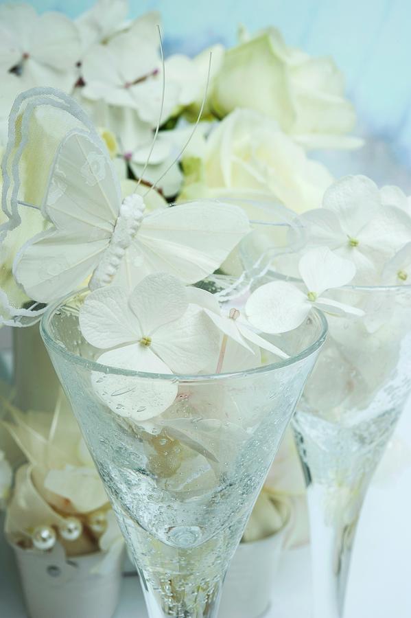 Stemmed Glasses Decorated With White Hydrangea Florets And Butterfly Ornaments In Front Of Hydrangea Plant Photograph by Linda Burgess