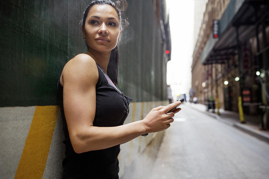 Architecture Photograph - Thoughtful Sporty Woman Holding Smart Phone While Leaning On Wall By Street by Cavan Images