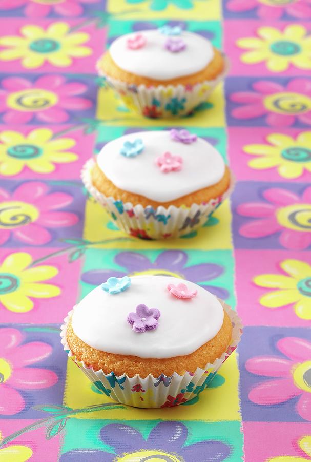 Three Fairy Cakes Sitting On A Coloured Flower Pattern Background Photograph by Stuart Macgregor