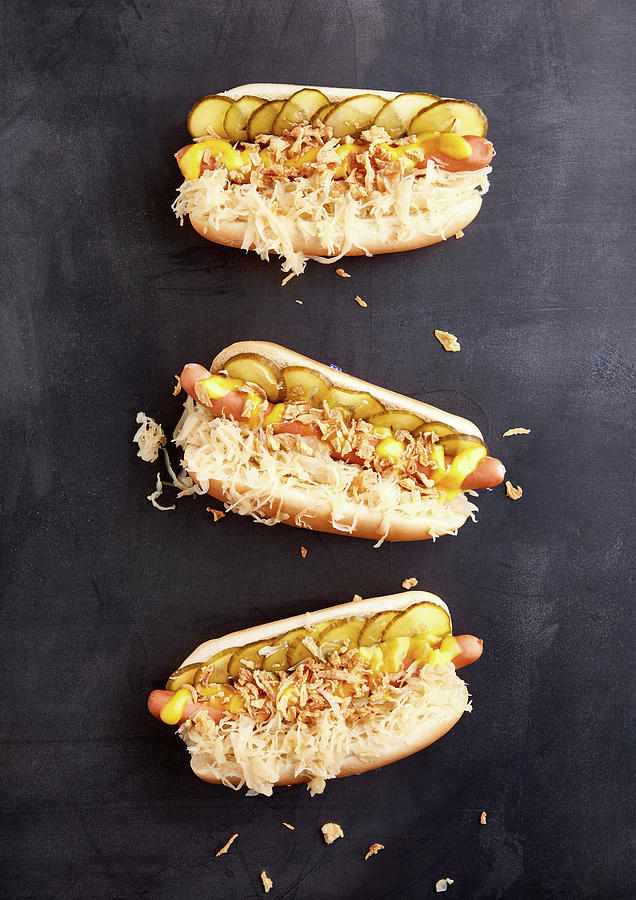 Three Hot Dogs With Cooked Sausages, Sauerkraut, Pickled Cucumbers, Mustard And Roasted Onions Photograph by Sylvia Meyborg