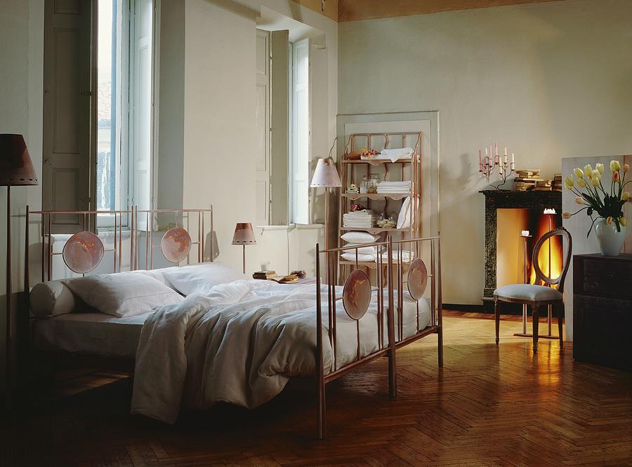 Traditional Bedroom With Tall Windows And Large Metal Bed With Painted Oval Panels; Firelight From Open Fireplace In Background Photograph by Laura Rizzi