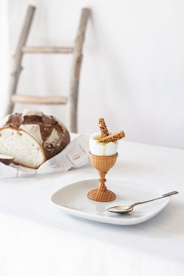 Easter Photograph - Two Crackers With Sesame Seeds In A Broken Boiled Egg On A Wooden Stand On A Table With A White Tablecloth by Cavan Images